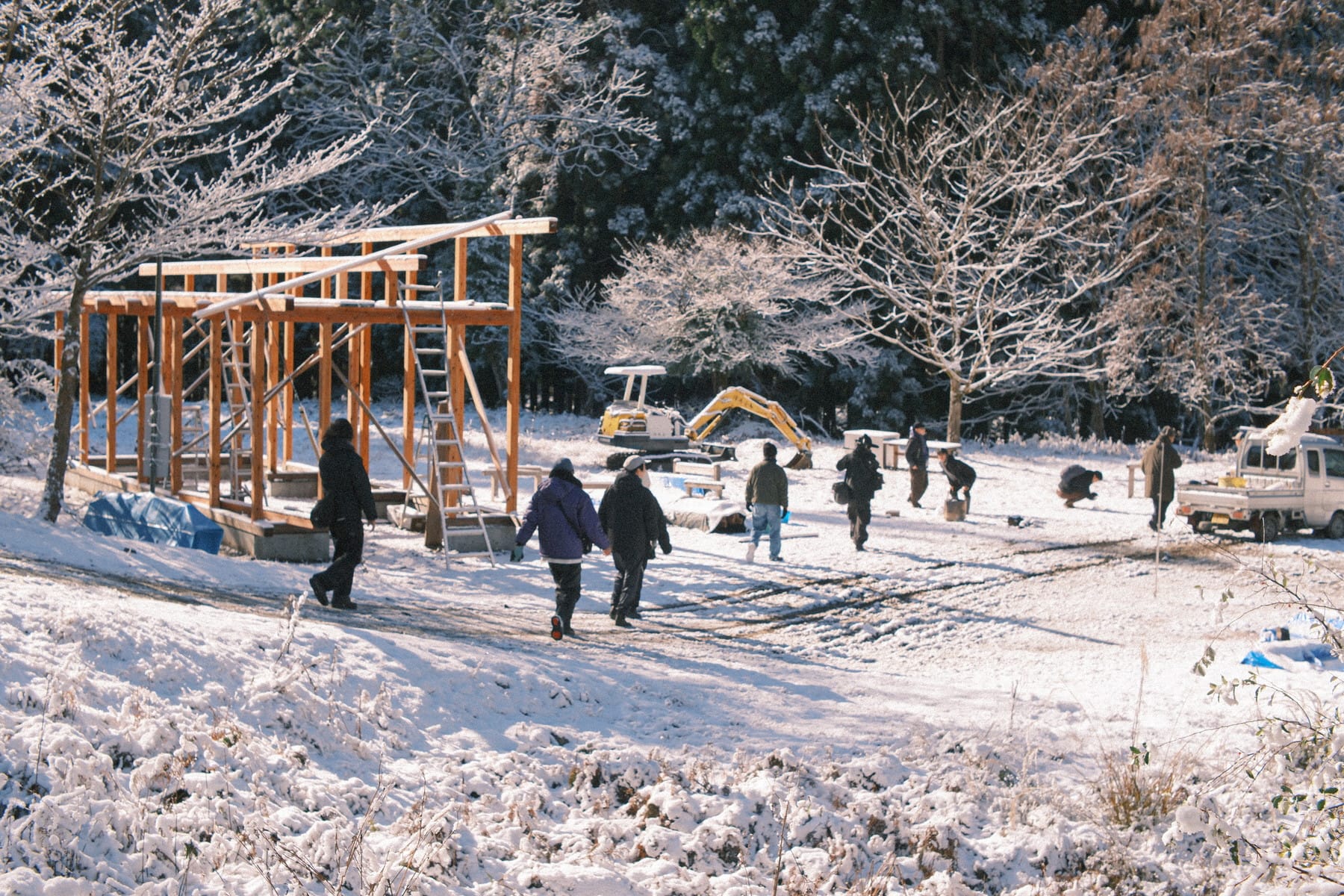 雪が積もる山林を歩く10名ほどの参加者。周囲にはトラックや重機が置かれている